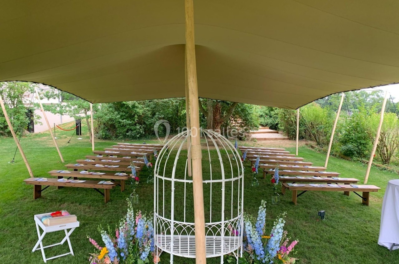 Chaises et bancs disposés sous une tente dans un jardin, avec une arche décorée de fleurs au premier plan.
