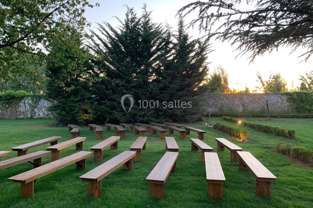Bancs en bois disposés en rangées sur une pelouse, entourés d'arbres et d'un mur en pierre au coucher du soleil.
