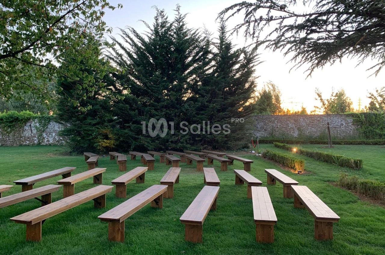 Bancs en bois disposés en rangées sur une pelouse, entourés d'arbres et d'un mur en pierre au coucher du soleil.