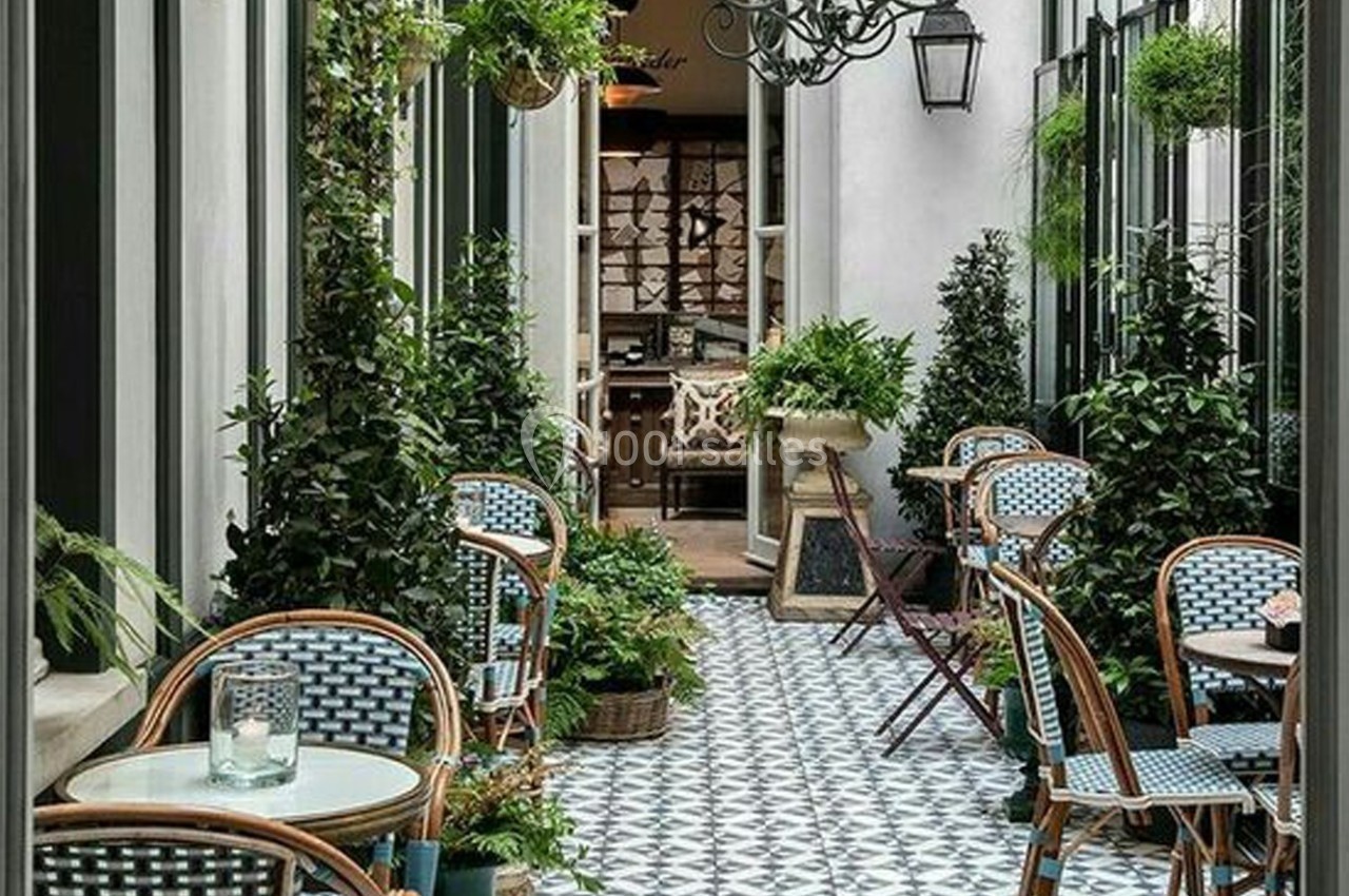 Terrasse étroite avec tables en rotin, chaises tressées, plantes vertes et sol carrelé à motifs géométriques.