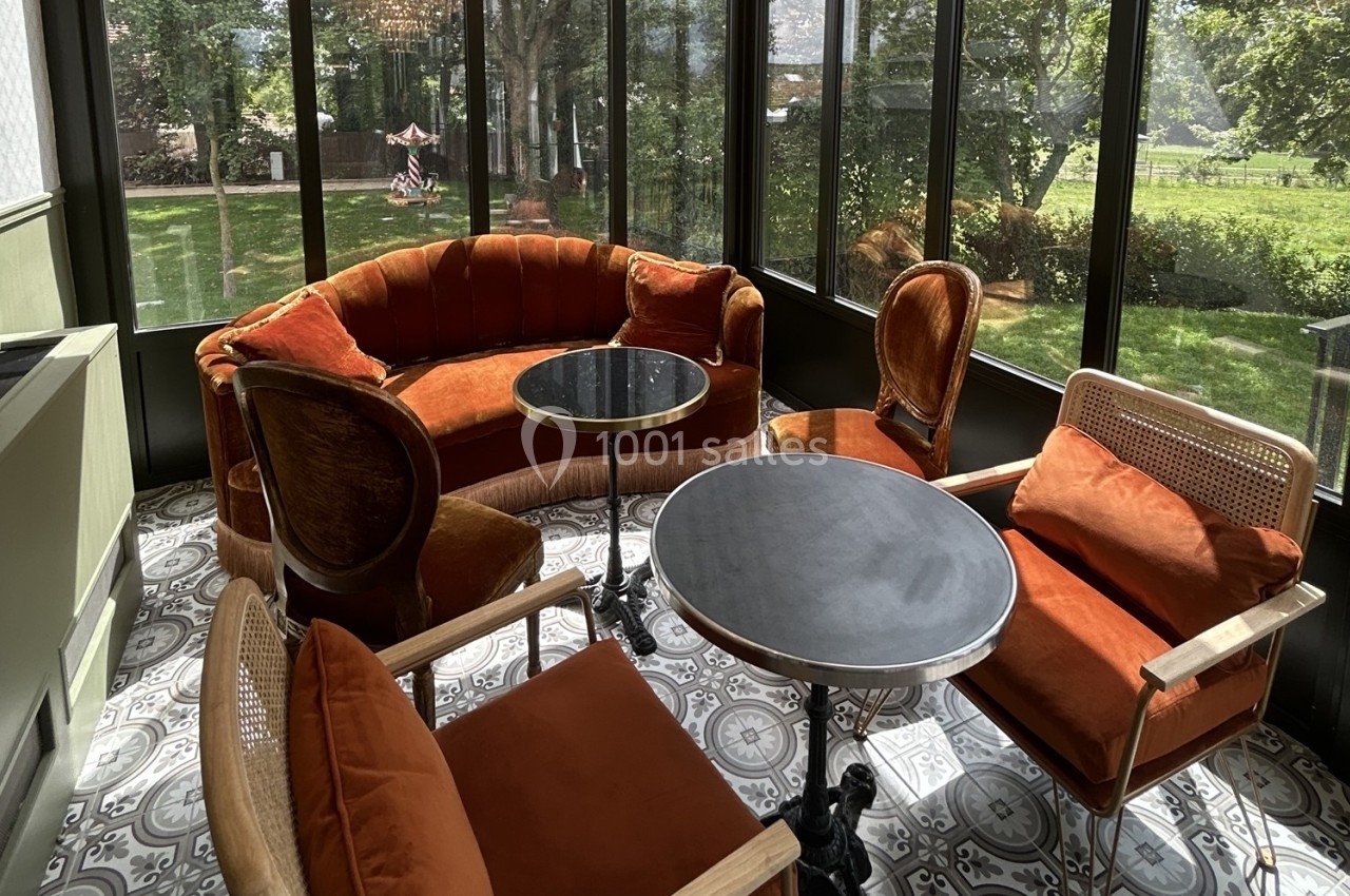 Salon lumineux avec fauteuils et canapé en velours orange, tables rondes et vue sur un jardin verdoyant.