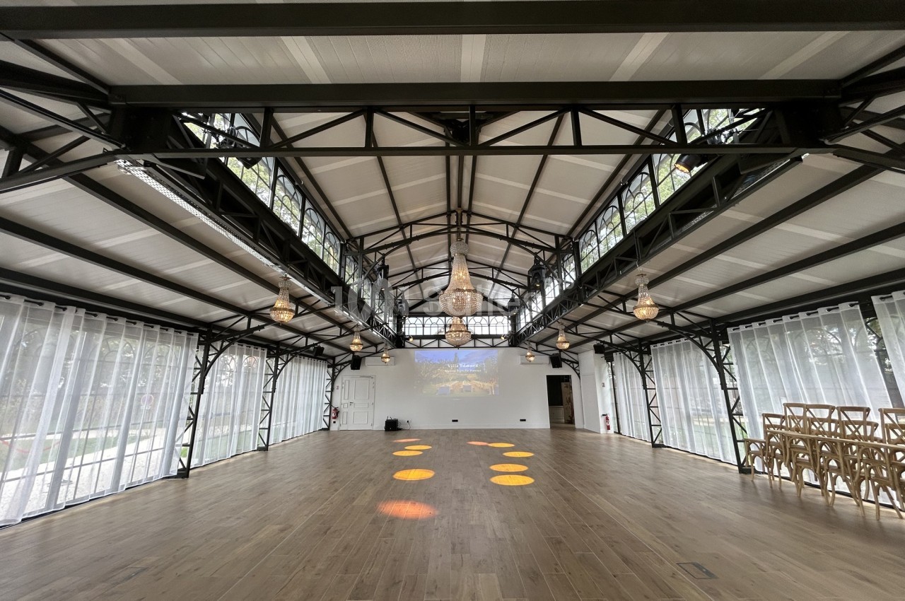 Grande salle lumineuse avec structure métallique, parquet en bois, baies vitrées et chaises alignées sur le côté.