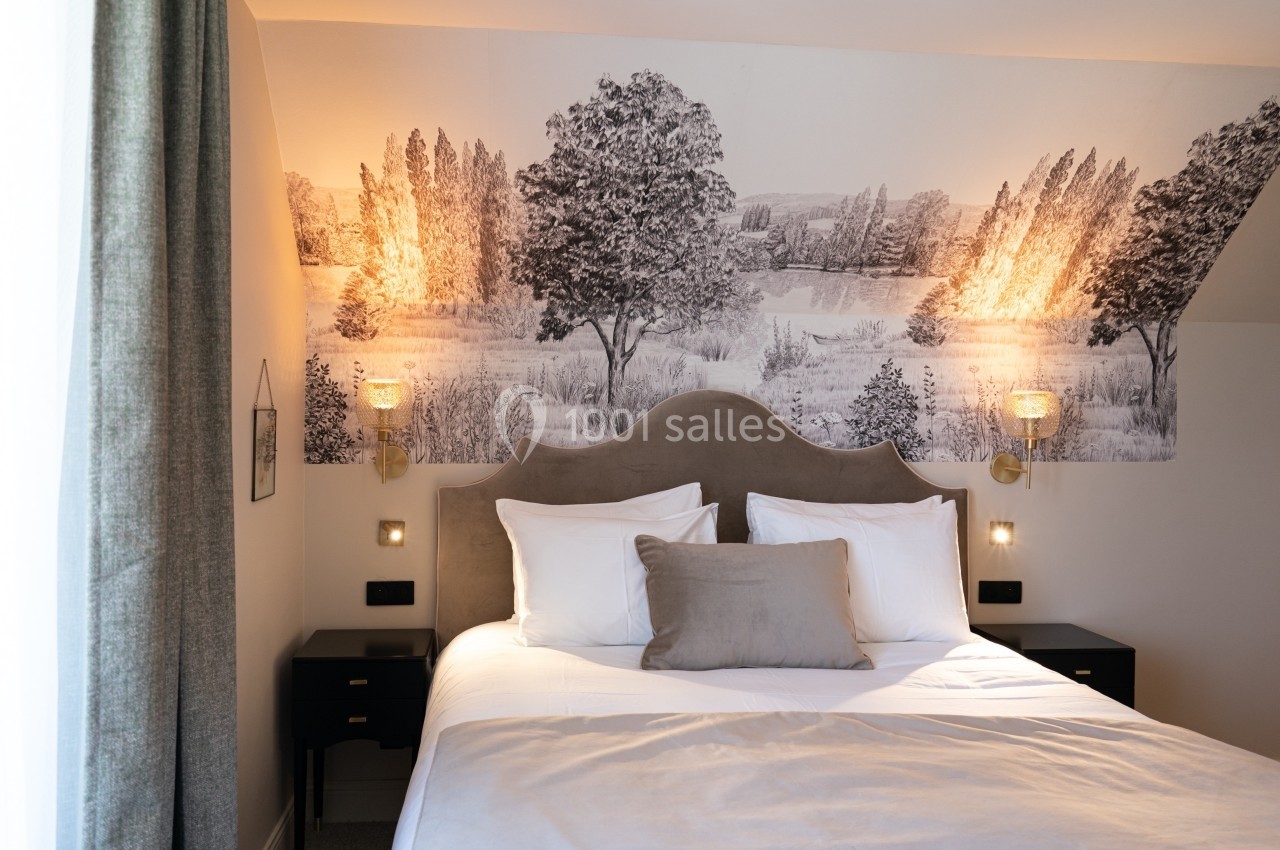 Lit double avec tête de lit grise, éclairé par deux appliques murales, devant un mur décoré d'une fresque paysagère.