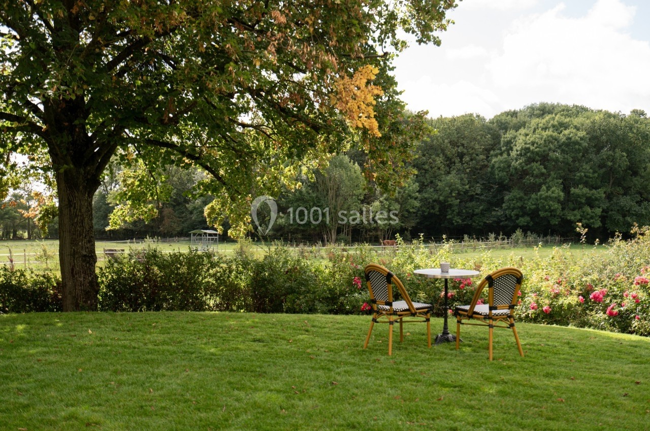 Table et deux chaises en plein air sur une pelouse, sous un arbre, avec vue sur un jardin et des champs verdoyants.