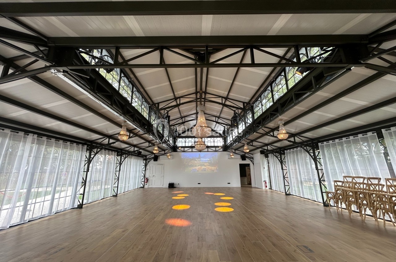 Salle lumineuse avec structure métallique, parquet en bois, lustres suspendus et chaises alignées sur le côté.