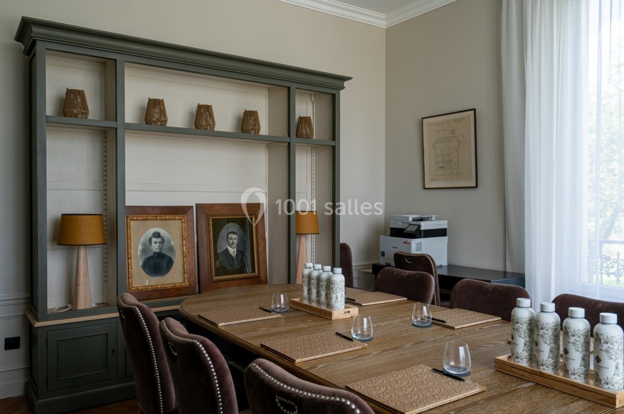 Salle de réunion avec table en bois, chaises, étagère décorative, portraits anciens et une imprimante en arrière-plan.