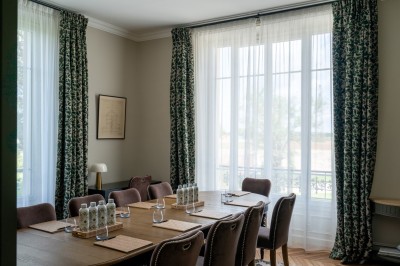 Salle de réunion lumineuse avec table en bois, chaises rembourrées, bouteilles d'eau et grandes fenêtres avec rideaux…
