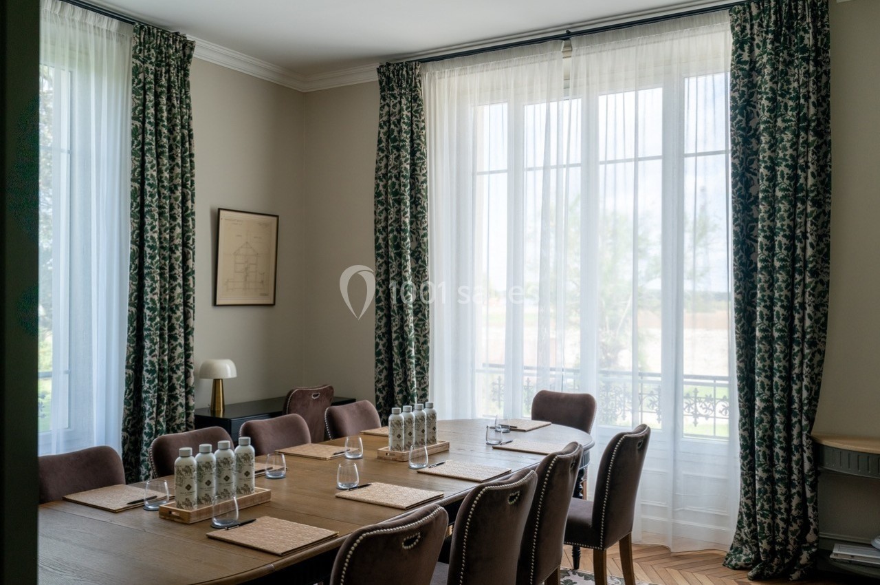 Salle de réunion lumineuse avec table en bois, chaises rembourrées, bouteilles d'eau et grandes fenêtres avec rideaux…