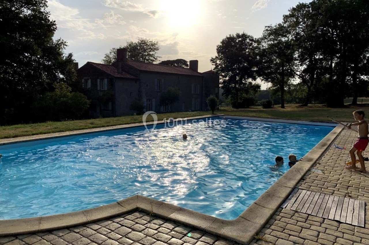 Piscine extérieure entourée de pavés, avec des enfants jouant dans l'eau près d'une maison en pierre au coucher du soleil.