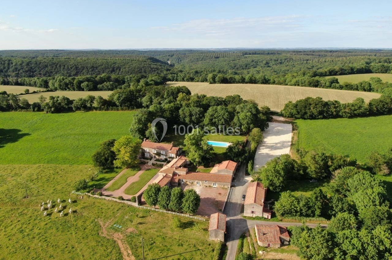 Vue aérienne d'une ferme avec bâtiments en pierre, piscine, champs verdoyants et forêt en arrière-plan.