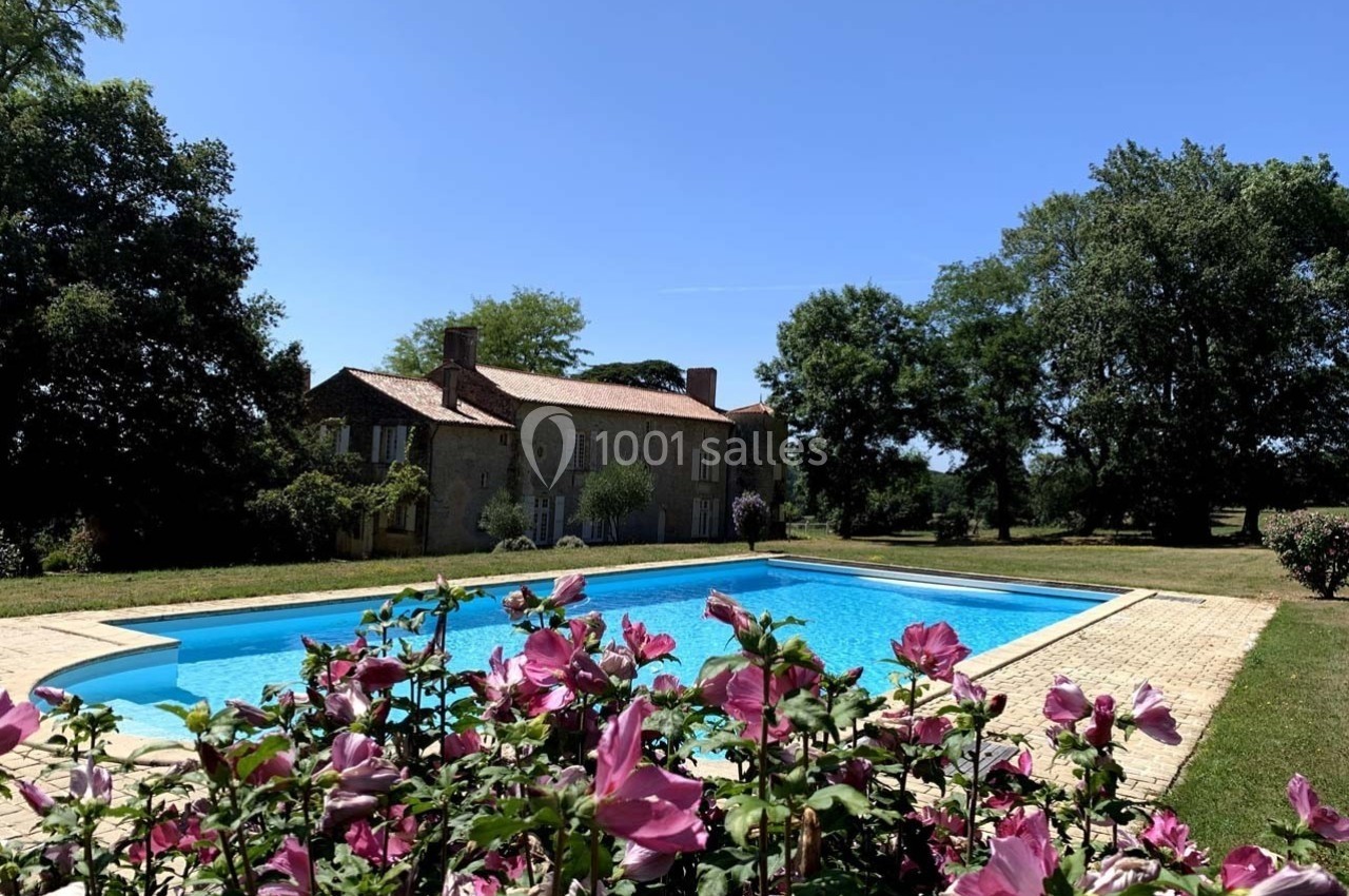 Maison en pierre entourée d'arbres avec une piscine au premier plan et des fleurs roses en avant-plan.