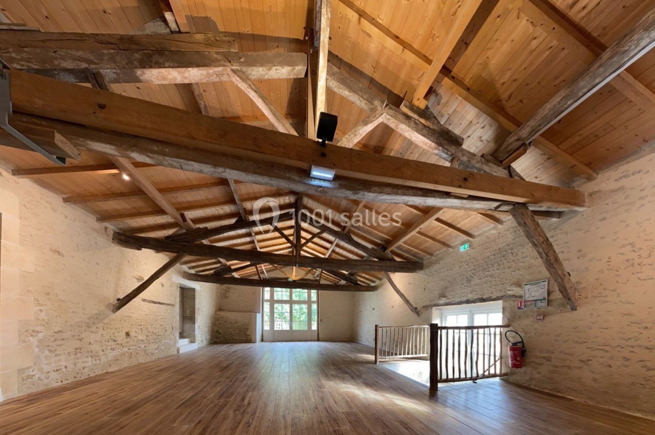 Salle spacieuse avec charpente en bois apparente, murs en pierre et parquet, éclairée par des fenêtres et des lumières…