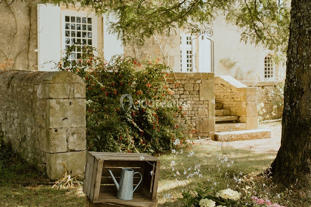 Arrosoir en métal posé sur une caisse en bois dans un jardin fleuri devant une maison en pierre.