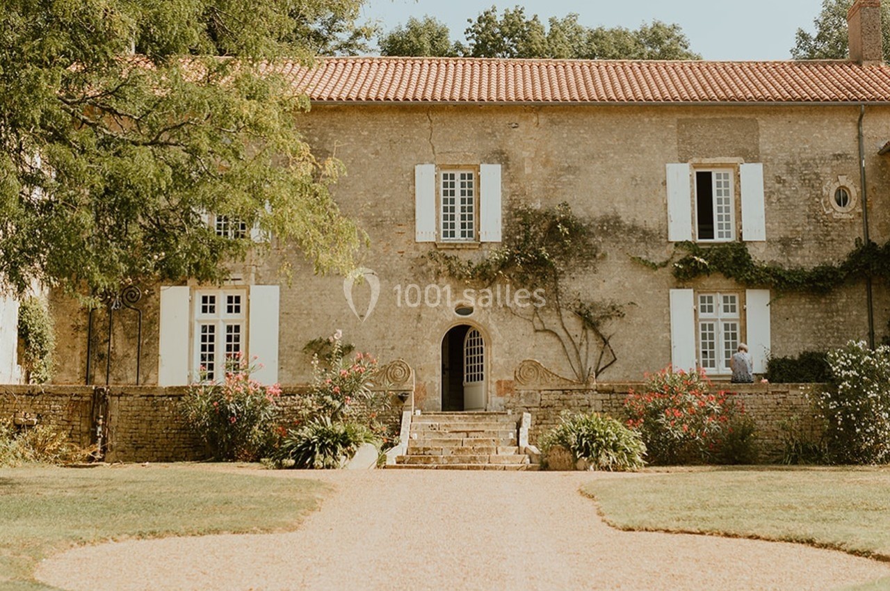 Façade d'une maison en pierre avec volets blancs, entourée de végétation et d'un jardin bien entretenu.