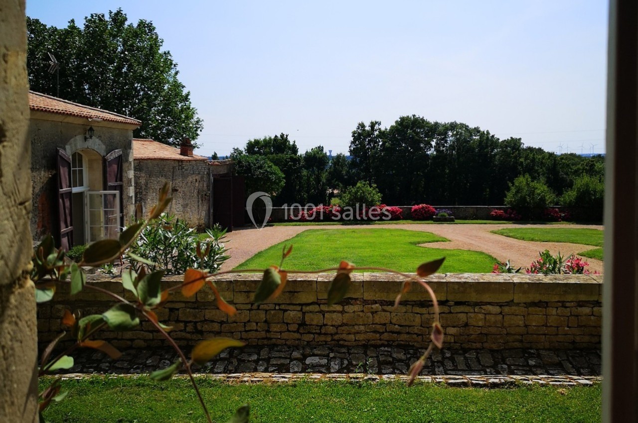 Vue sur un jardin avec pelouse, allées gravillonnées, massifs fleuris et bâtiments en pierre sous un ciel dégagé.