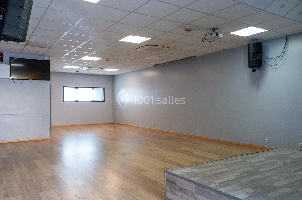 Salle vide avec parquet, murs gris, plafond à dalles blanches, éclairage intégré et petite estrade au premier plan.