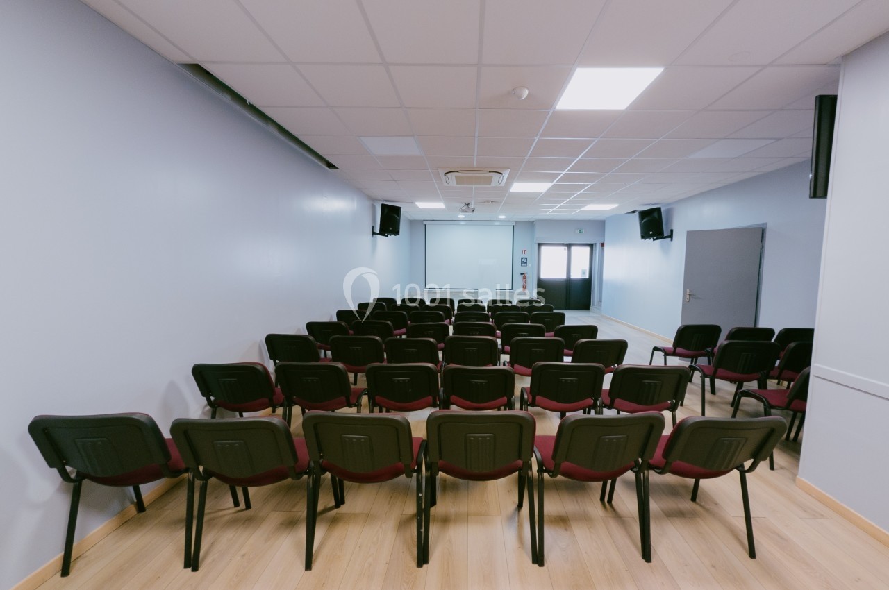 Salle de conférence vide avec des rangées de chaises rouges et noires, un écran de projection et un sol en bois clair.