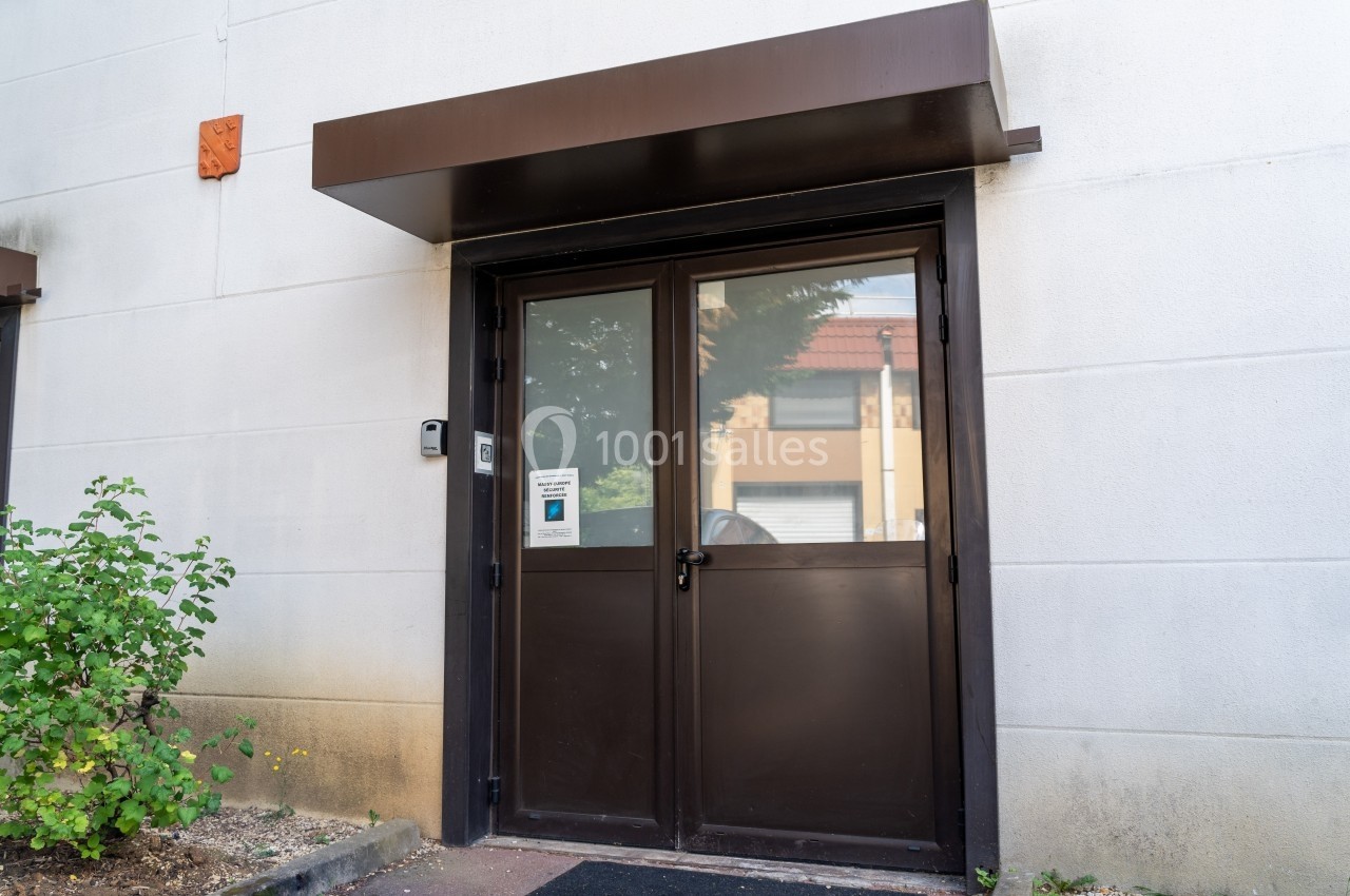 Porte d'entrée vitrée en métal sombre avec auvent, interphone et végétation sur le côté gauche.