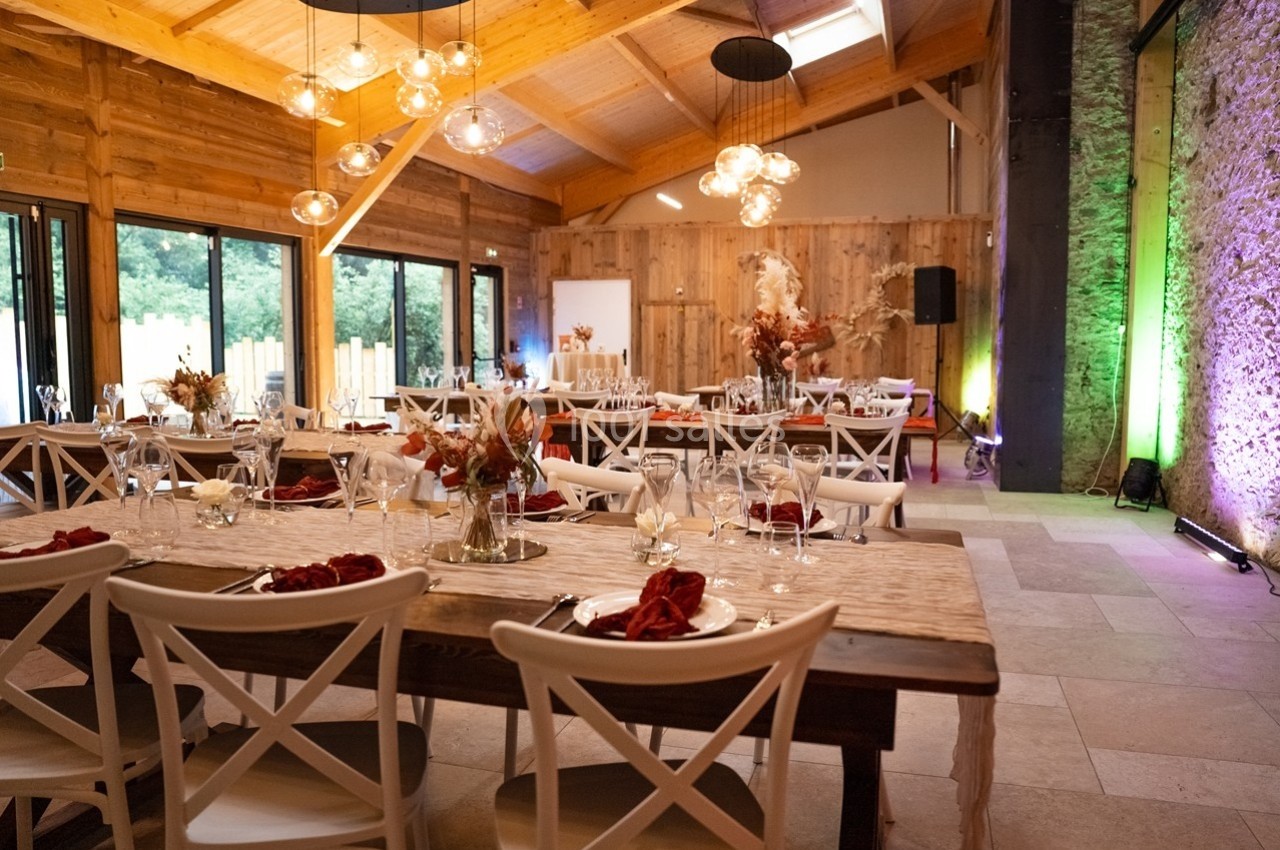 Salle de réception en bois avec tables décorées, chaises blanches et lumière naturelle entrant par de grandes baies vitrées.