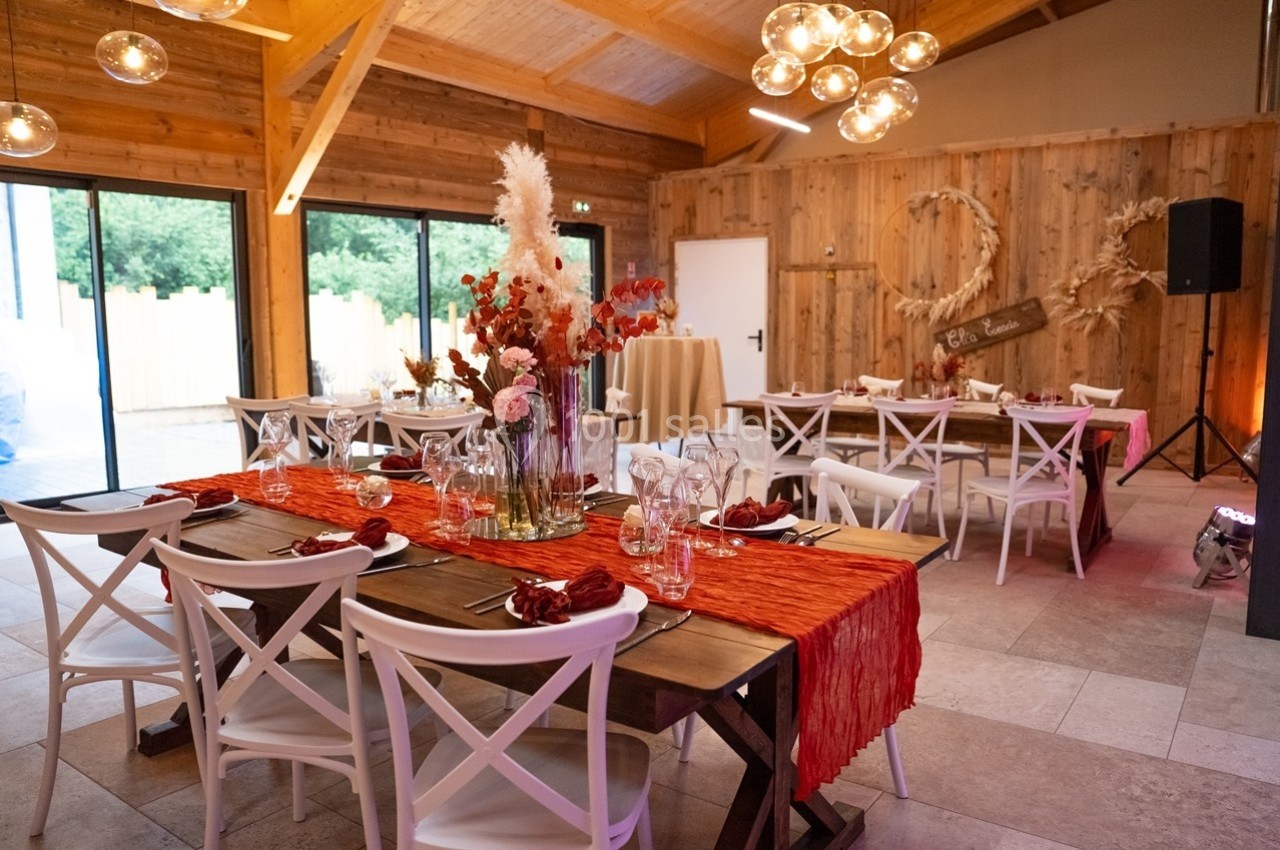 Salle décorée pour un événement, avec tables en bois, chaises blanches, nappes rouges et ornements floraux.