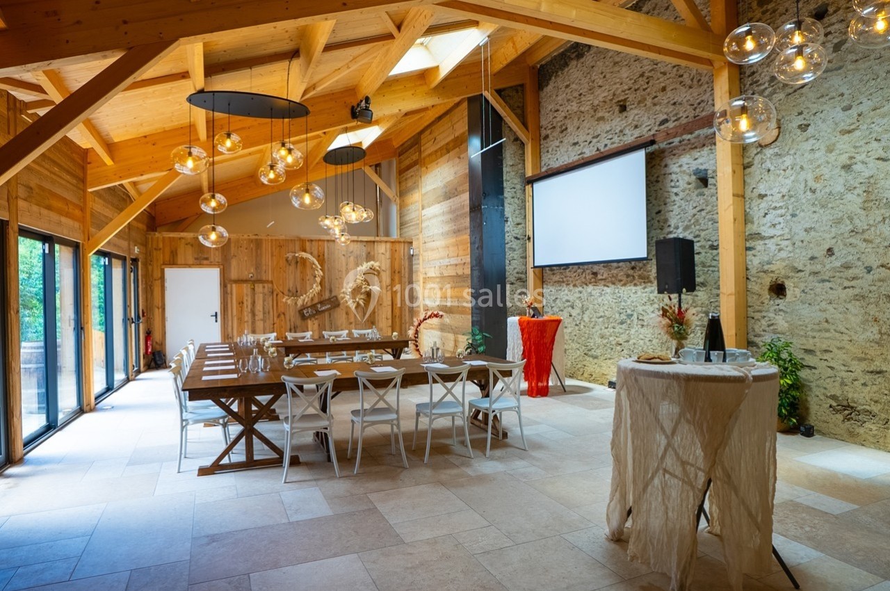 Salle de réception lumineuse avec poutres apparentes, table dressée, écran de projection et décorations modernes.