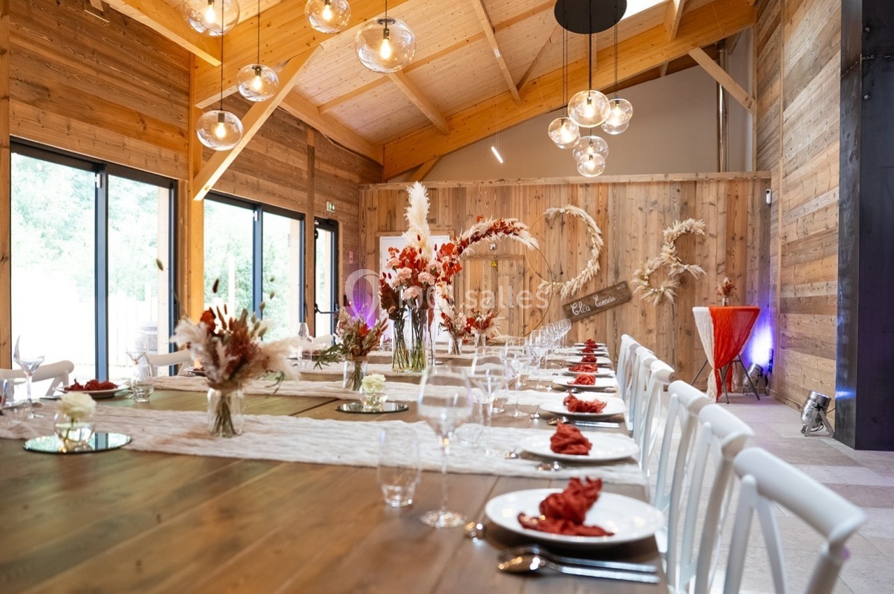 Salle de réception en bois avec tables décorées de fleurs séchées, serviettes rouges et grandes baies vitrées.