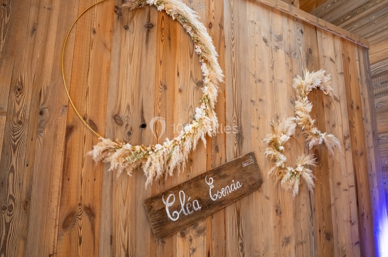 Anneaux décoratifs en fleurs séchées et panneau en bois gravé ’Cléa & Enora’ accrochés sur un mur en bois.