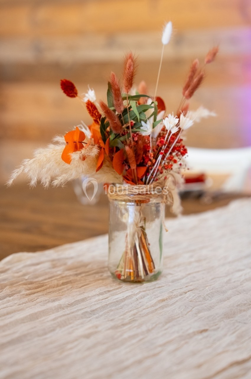 Bouquet de fleurs séchées aux tons rouges et orangés dans un pot en verre sur une nappe beige.