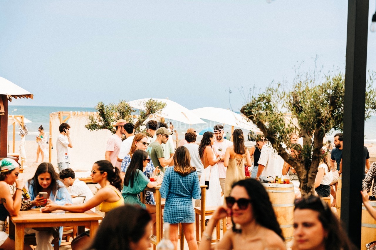 Groupe de personnes discutant et se détendant autour de tables en bois sur une plage ensoleillée.