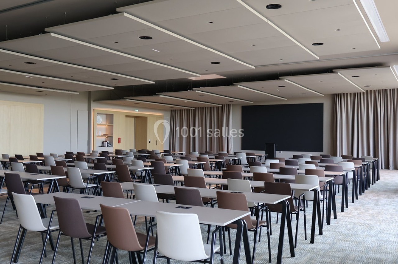 Salle de conférence moderne avec des rangées de tables et de chaises vides, écran noir et rideaux en arrière-plan.