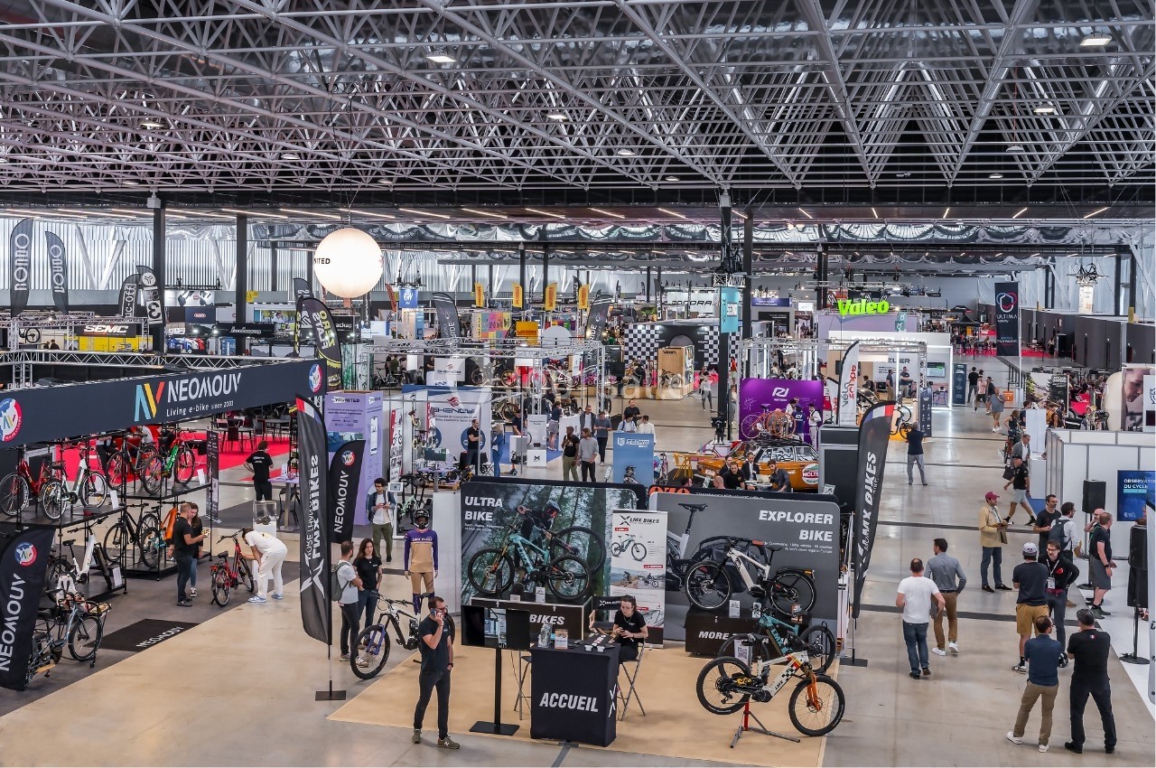 Vue d'un salon professionnel avec des stands exposant des vélos et des accessoires dans un grand hall lumineux.