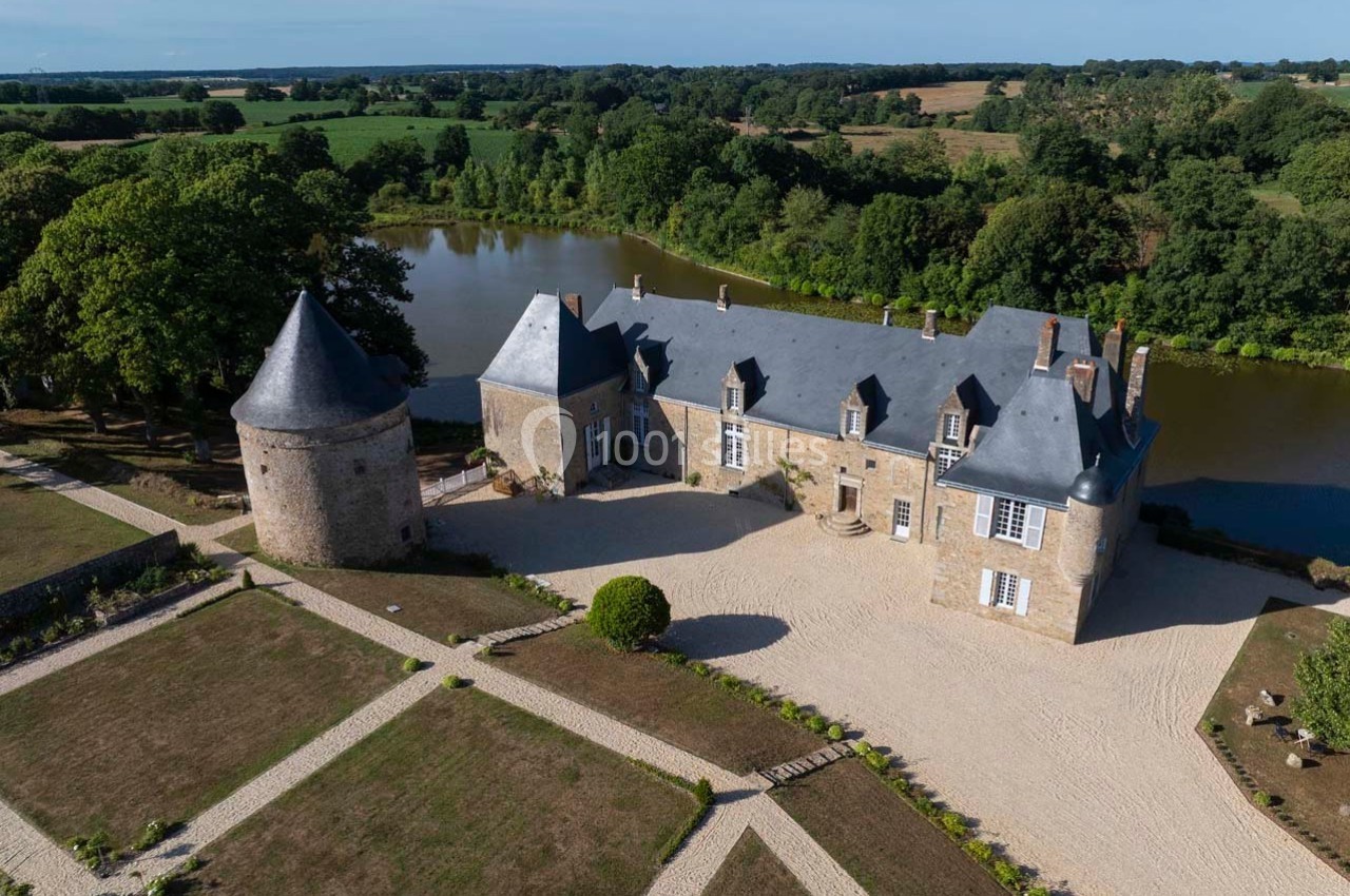 Vue aérienne d'un château en pierre avec une tour ronde, entouré d'un parc, d'un étang et de paysages verdoyants.