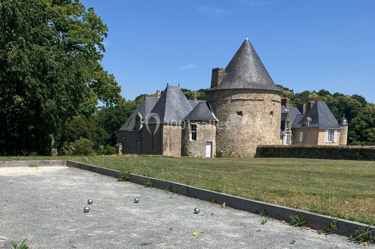 Terrain de pétanque devant un château en pierre avec tours et toits en ardoise, entouré de verdure.