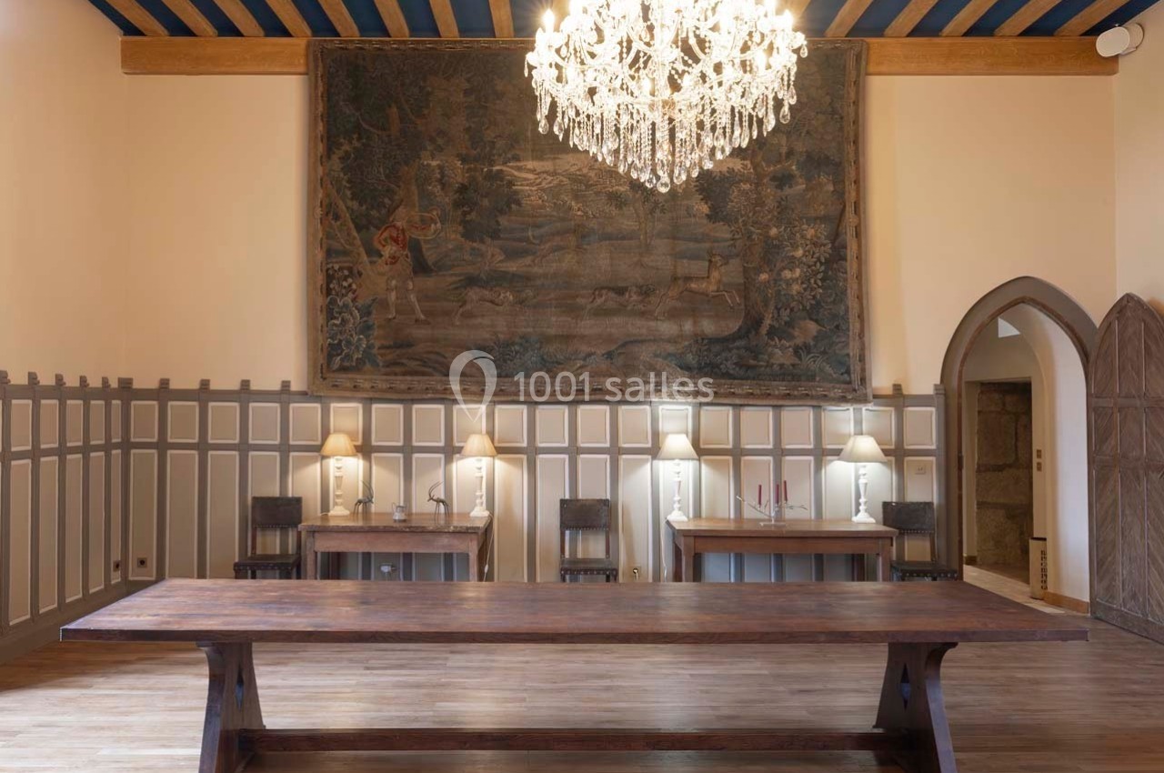 Salle avec une grande table en bois, lustre en cristal, tapisseries murales et éclairage doux par des lampes de bureau.
