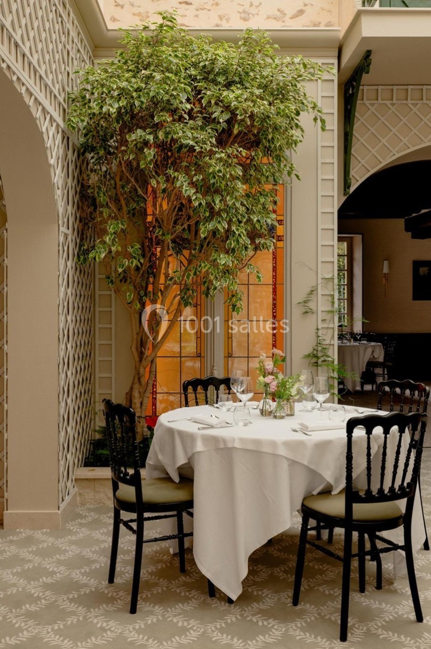 Table dressée avec nappe blanche, verres et couverts, entourée de chaises noires dans une salle lumineuse avec un arbre…