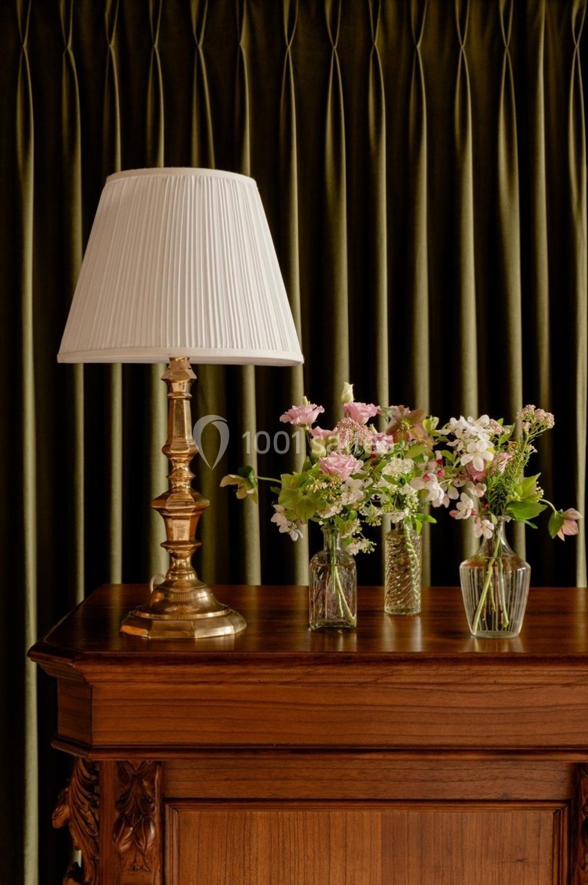 Lampe à abat-jour blanc sur un meuble en bois, accompagnée de petits vases contenant des fleurs, devant un rideau vert.