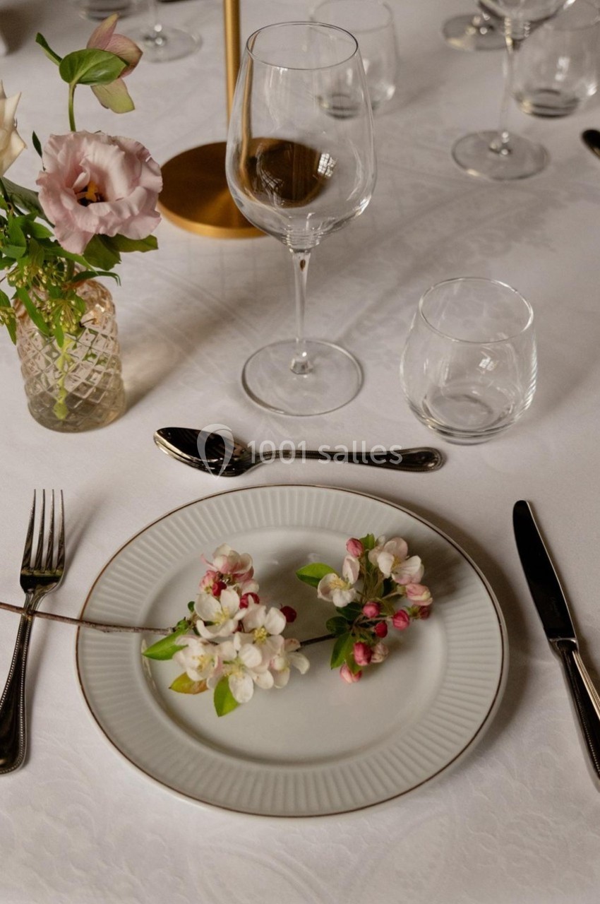 Assiette blanche décorée de branches fleuries sur une table dressée avec verres, couverts et vase de fleurs.