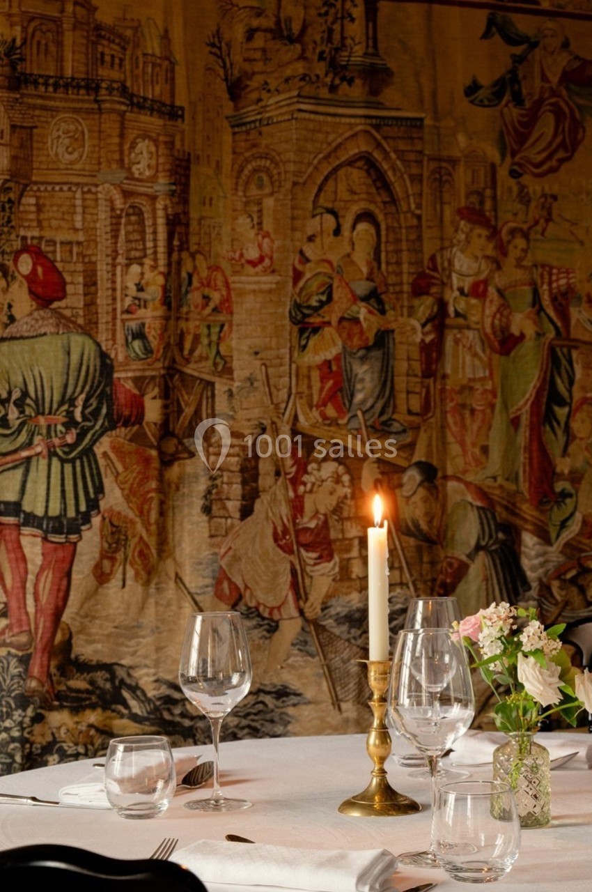 Table dressée avec bougeoir allumé, verres et fleurs, devant une tapisserie médiévale représentant une scène historique.