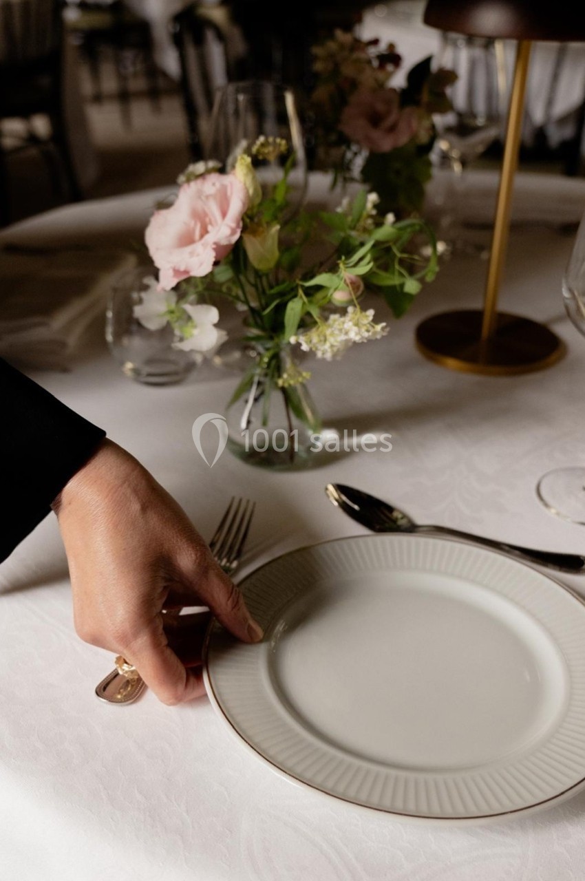 Main plaçant une assiette blanche sur une table dressée avec des fleurs, des couverts et des verres.
