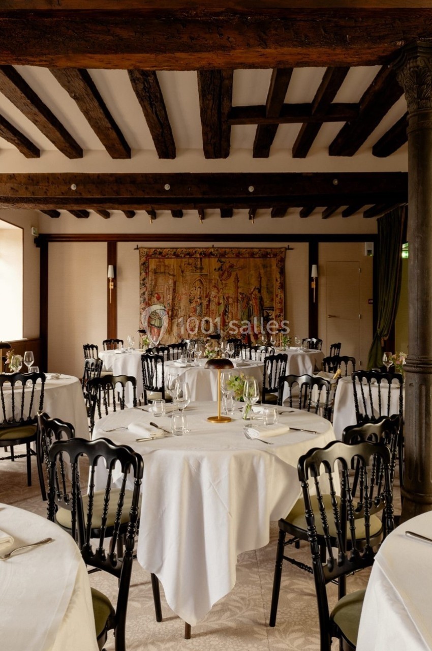 Salle de restaurant élégante avec tables rondes dressées, poutres apparentes et tapisserie murale décorative.