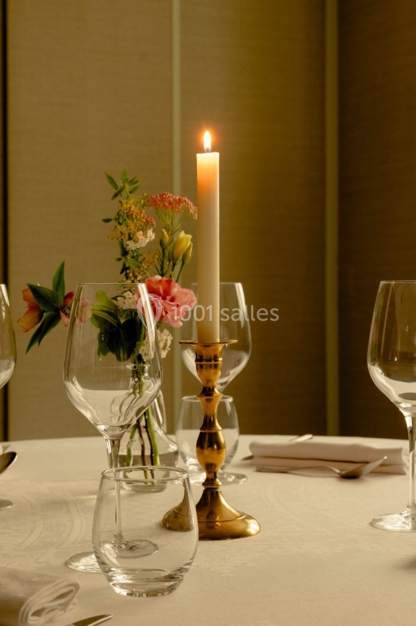 Bougie allumée sur un chandelier doré au centre d'une table dressée avec verres, fleurs et serviettes.