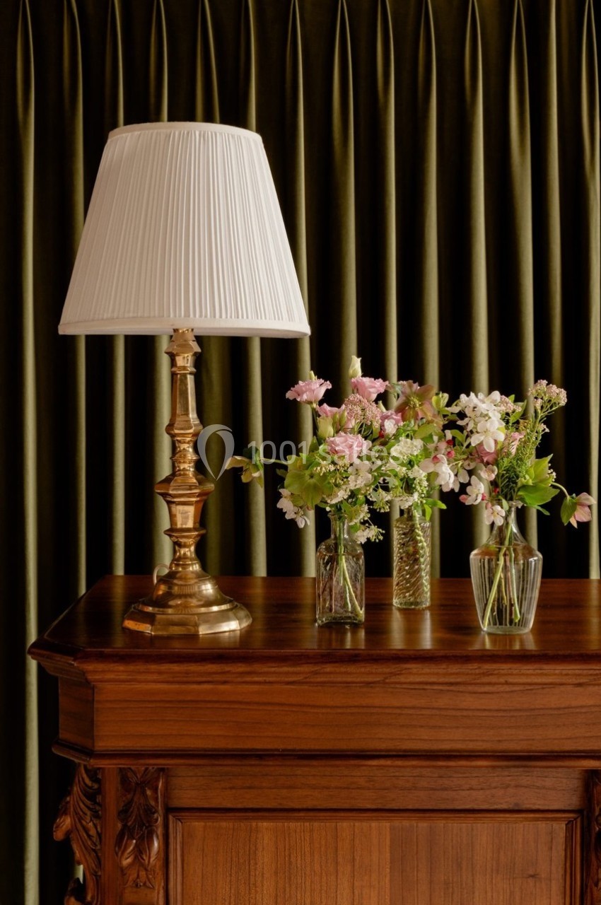 Lampe à abat-jour blanc et bouquets de fleurs sur un meuble en bois devant des rideaux verts.