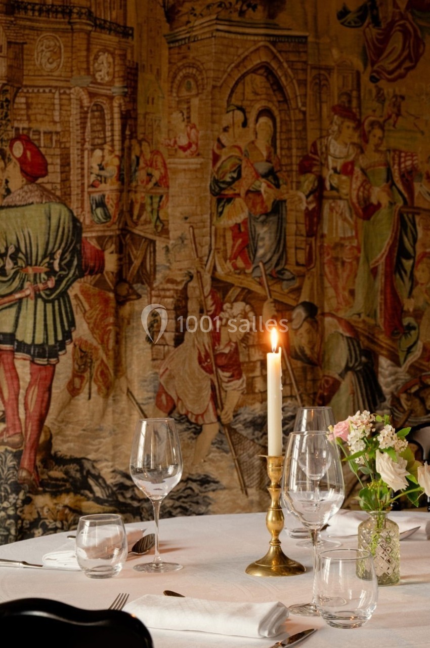 Table dressée avec élégance, bougie allumée et décor mural représentant une scène médiévale.