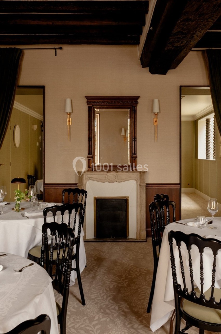 Salle de restaurant élégante avec tables dressées, cheminée décorative et poutres apparentes au plafond.