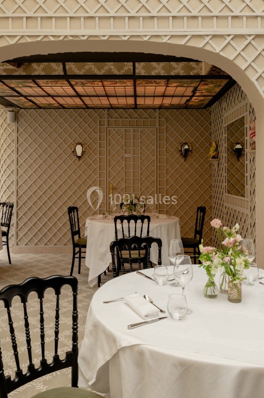 Salle de restaurant élégante avec tables dressées, chaises noires et décoration murale en treillis beige.
