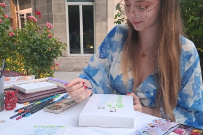 Illustration d'une femme souriante en tenue décontractée, posée sur une table en bois avec des coquillages en arrière-plan.