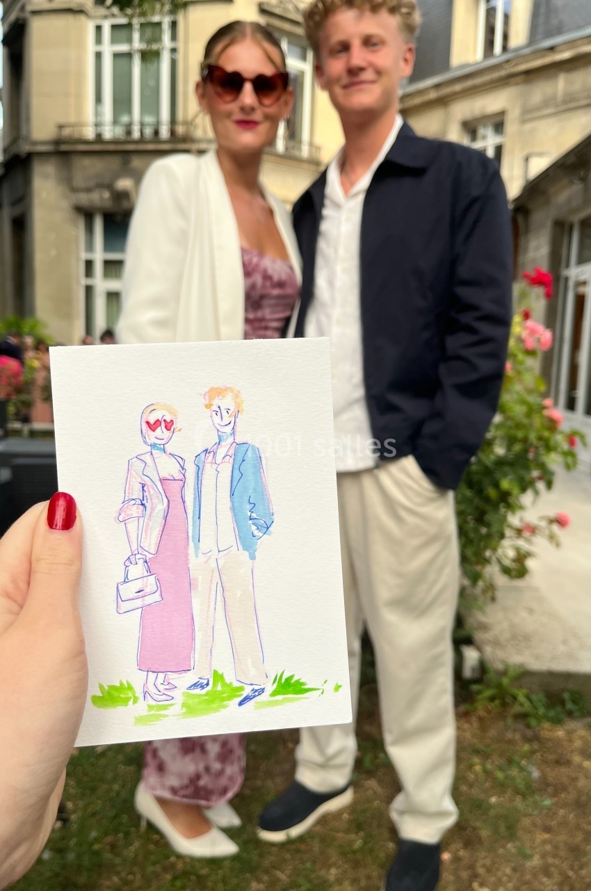 Une main tient un dessin représentant un couple élégant, avec le couple réel en arrière-plan dans un jardin.