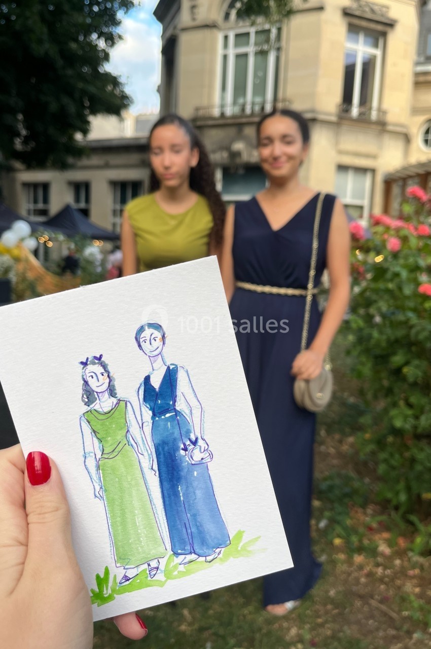 Une main tient un dessin représentant deux femmes posant devant un jardin fleuri et des bâtiments en arrière-plan.