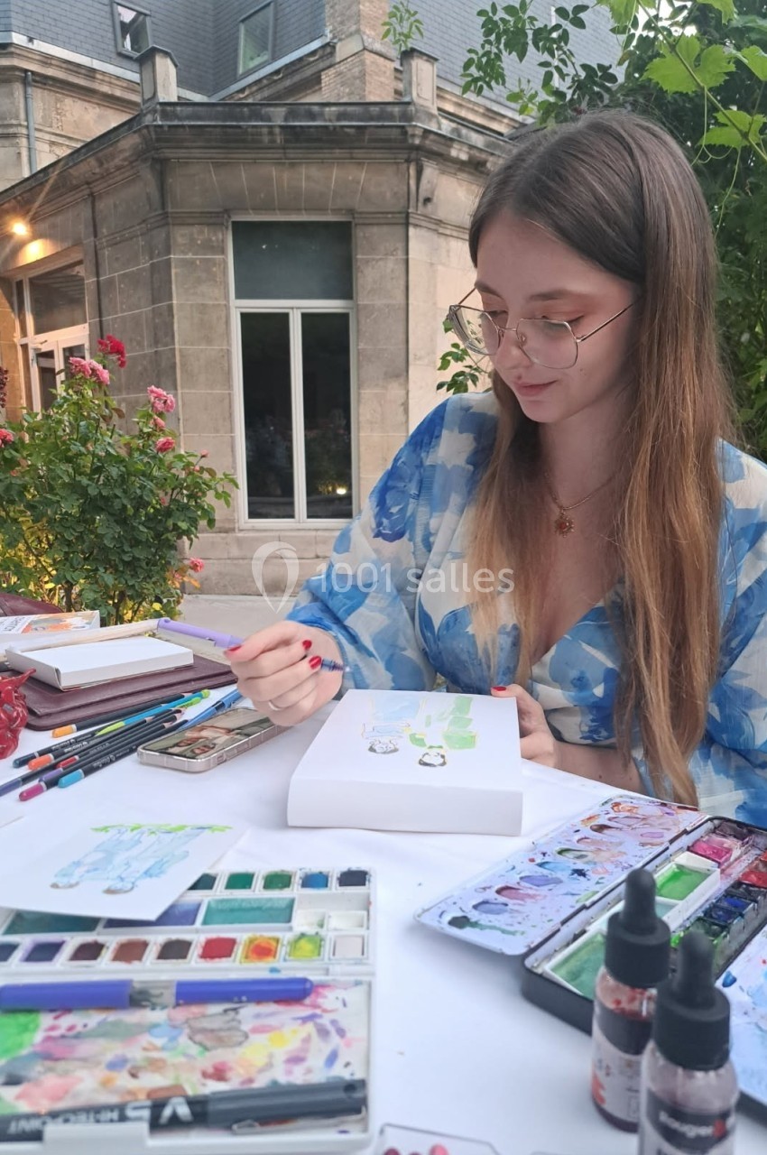 Une femme peint à l'aquarelle sur une table en extérieur, entourée de fournitures artistiques et de végétation.