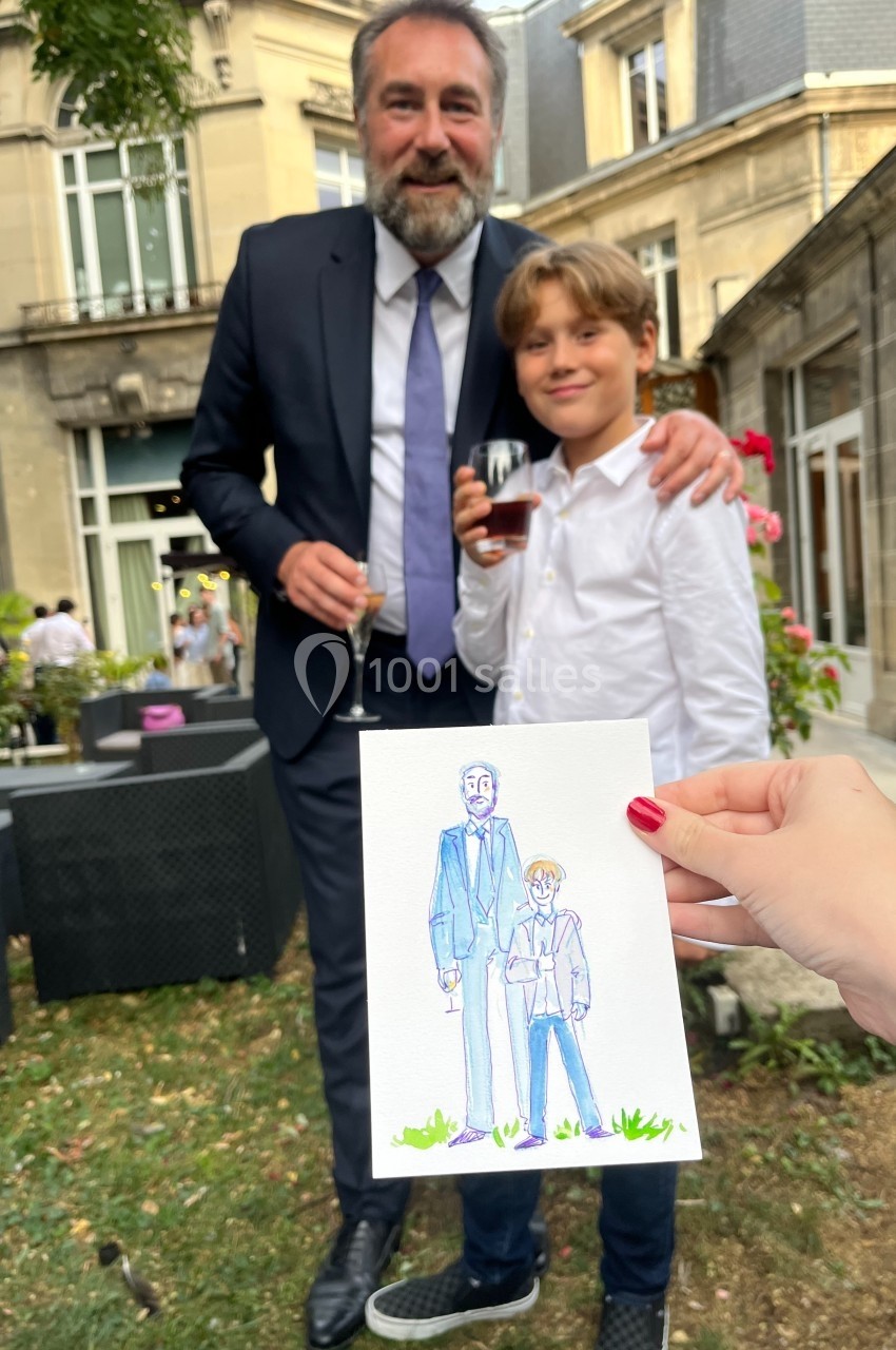 Un homme en costume et un enfant souriant posent dans un jardin, avec un dessin les représentant tenu au premier plan.